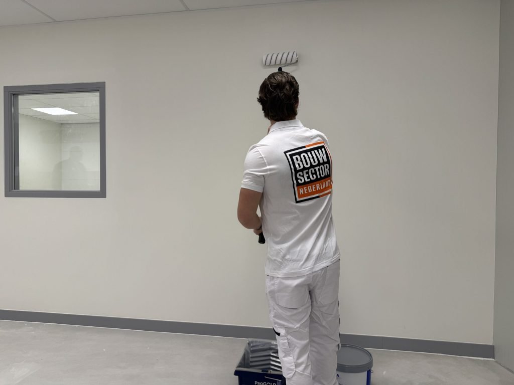 Schilder van Bouwsector Nederland saust een lichte muur naast een binnenraam, met verfbak en emmer op de vloer
