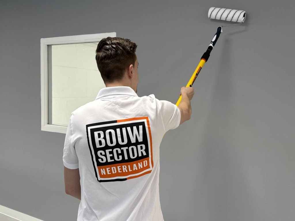 Schilder van Bouwsector Nederland rolt grijze muurverf op een wand naast een raam met witte omlijsting