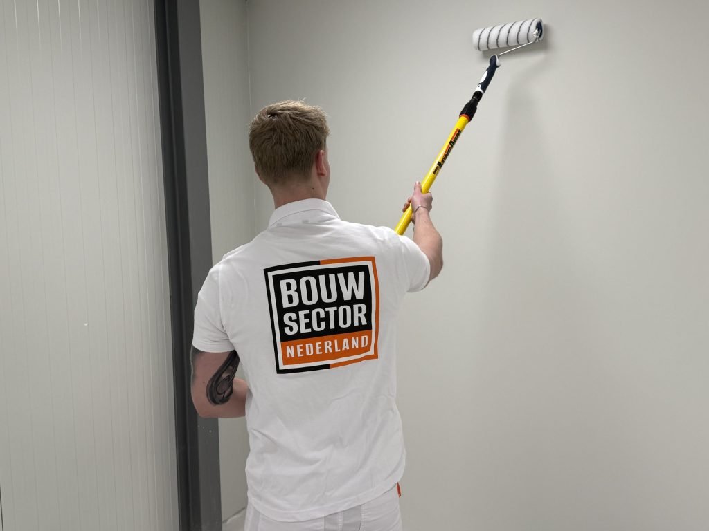 Schilder met logo van Bouwsector Nederland rolt een lichte wand bij een deurpost in een smalle ruimte